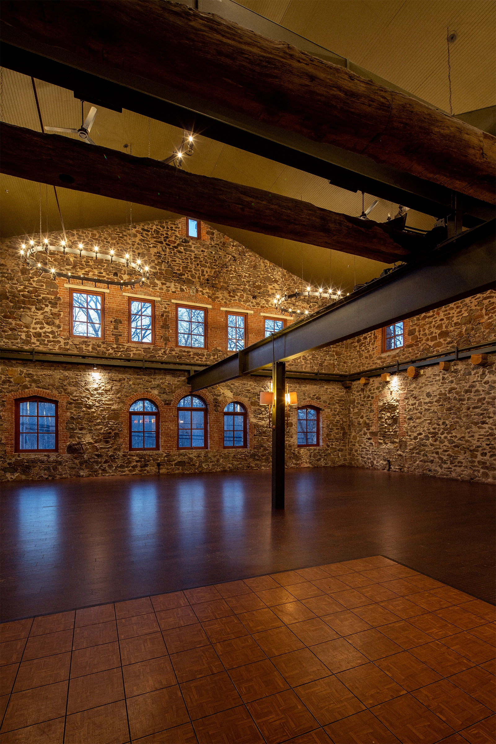 Empty Grand Salon with fieldstone walls, arched windows, timber beams, chandeliers, and wood floor.