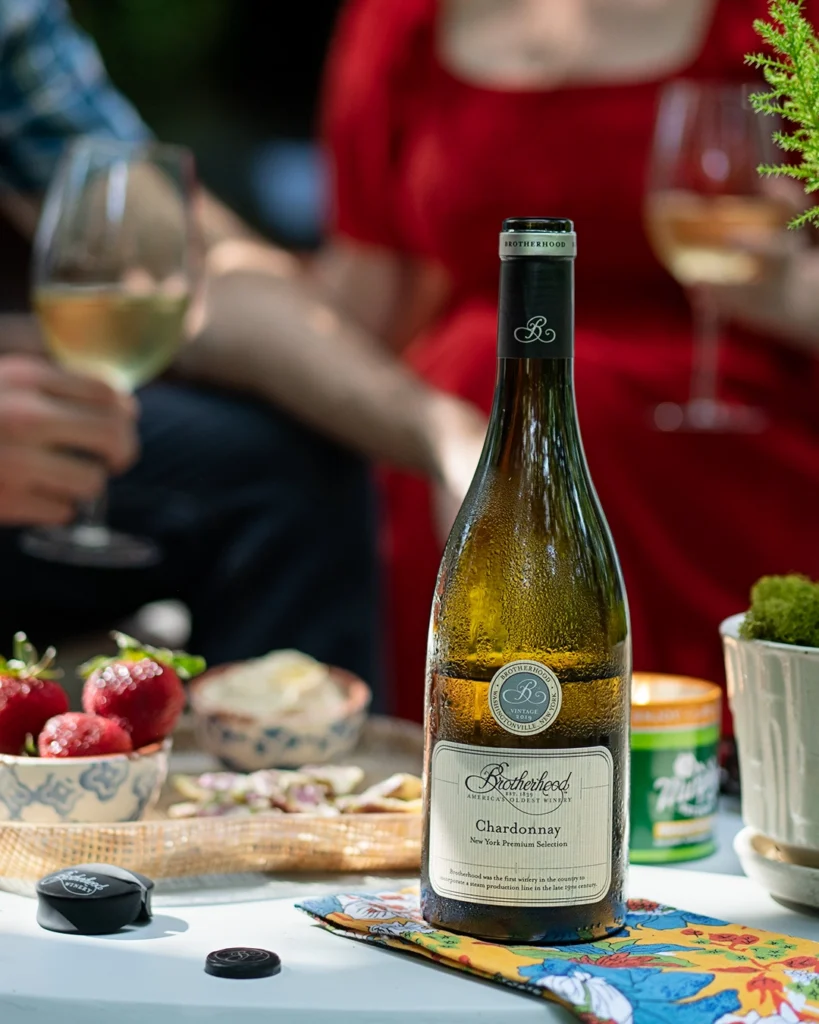 Chardonnay bottle on outdoor table with strawberries and wine glasses, people seated in background